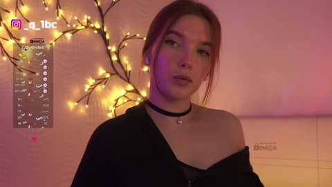 Video of a young woman with fair skin and brown hair, wearing a black choker and off-shoulder top, standing in a dimly lit room with warm, yellow fairy lights and a white wall.