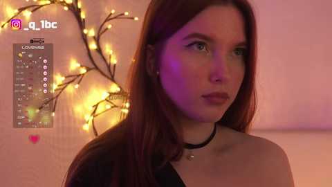 A video of a young woman with long brown hair, wearing a black choker and black top, standing against a soft pink background with warm, glowing fairy lights.