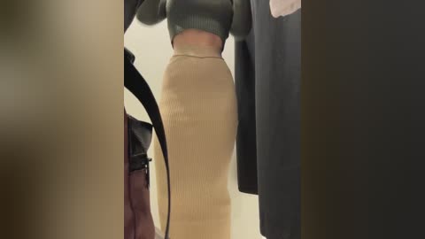 Video of a person wearing a fitted, ribbed beige skirt and black sweater, carrying a black handbag, standing in a dimly lit, neutral-toned room.