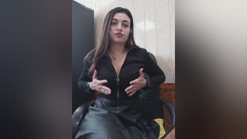 Media: Video of a Latina woman with long dark hair, wearing a black leather jacket and black leather skirt, seated in a black office chair, in a dimly lit room with beige walls.