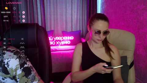 Video of a young, slender Caucasian woman with long brown hair in a ponytail, wearing sunglasses, a black sleeveless dress, and a beige chair. She's engrossed in her phone. Background: neon sign, plush couch, and colorful decor.