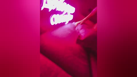 A video featuring a partially obscured nude woman with fair skin, lying on a red surface. The image is bathed in vibrant pink lighting, with white text partially visible in the background.