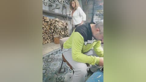 Media: Video of a man in a green shirt, seated on a chair, with a woman in a white top and jeans standing behind him. Background features a woodpile and decorative metal items.