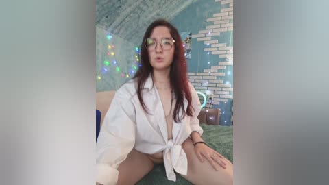 Video of a young woman with long, dark hair, fair skin, and large glasses, wearing an untied white shirt, sitting on a green carpet in a room with teal walls and brick pattern, string lights, and a plush toy.