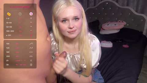 Media: Video of a young, blonde-haired, fair-skinned woman in a white blouse, holding an erect penis in her hand, in a bedroom with a bed and pink plush toys.