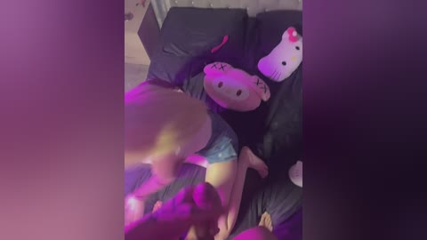 Media: A video of a person with light skin and long black hair lying on a black bed, wearing blue shorts, surrounded by Hello Kitty plushies, with a pink glow.