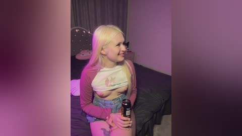 Media: Video of a blonde woman with pale skin, wearing a pink long-sleeve shirt lifted to reveal a small breast, denim shorts, and holding a can. Background shows a bed with dark bedding, pink lighting, and a mirror.