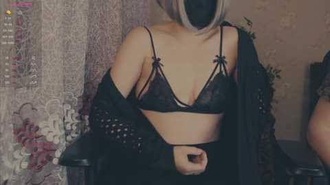 Video of a fair-skinned woman with short blonde hair, wearing a black lace bra and matching robe, sitting indoors, partially obscured by a lace curtain.