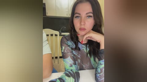 Media: Video of a fair-skinned woman with long, wavy brunette hair, wearing a sheer, multicolored floral top, sitting in a modern kitchen with white cabinets and black countertops.