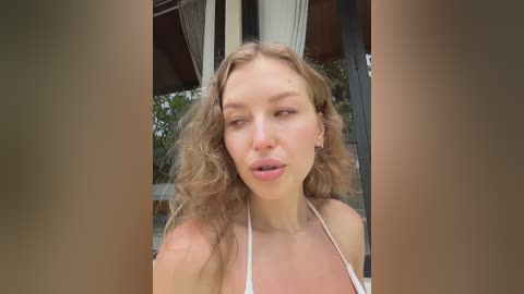 Media: Video of a young, fair-skinned woman with curly blonde hair, wearing a white bikini top, smiling and making a kissy face outdoors near a modern building with large glass windows.