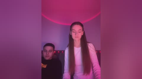 Media: Video of a young woman with long brown hair, wearing a white long-sleeve shirt, standing in a dimly lit room with a red hue. A boy with glasses and a black hoodie is seated behind her.