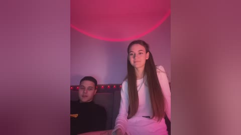 Media: Video of a young woman with long brown hair, wearing a white top, standing in front of a seated young man with short black hair, both under a pinkish-red light in a dimly lit room.