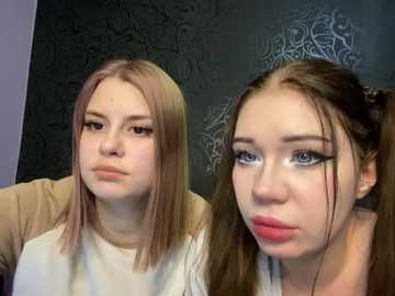 Media: Video of two young women with fair skin, one with pink hair and the other with long brown hair styled in pigtails, both wearing white tops, against a dark patterned background.