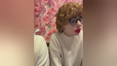 Media: Video of a fair-skinned person with short, curly red hair, wearing black-rimmed glasses and bright red lipstick, sitting in a room with floral-patterned wallpaper.