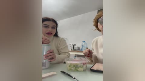 Media: A video of two women, one with dark hair and the other with light hair, sitting at a white table in a modern, bright room. The woman with dark hair holds a cup, and the other holds a clear container with a salad.