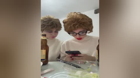 Media: Video of two women with short, curly brown hair, wearing glasses, white turtlenecks, and red lipstick, sitting at a table, one holding a beer bottle, the other a smartphone.