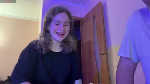 Media: Video of a young woman with curly brown hair, wearing a black top, laughing joyfully, standing in a doorway next to a man in a light blue shirt.