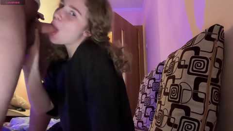 Media: A video captures a young woman with curly hair, wearing a black top, performing oral sex on a nude man, seen from the side, in a bedroom with a patterned pillow.