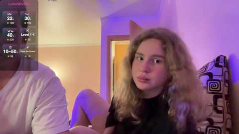 Media: A video of a young woman with curly blonde hair, wearing a black top, sitting on a bed with a patterned pillow in a dimly lit room. The background shows a wall and a partially open door.
