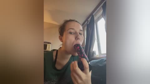 A video of a young woman with light skin, dark hair, and a green top, eating a black-and-red lollipop, standing near a window with curtains, in a dimly lit room.