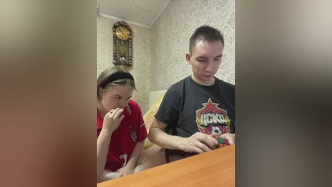 A video shows a young man and woman, both Caucasian, in a small, dimly lit room with patterned wallpaper. The man wears a black T-shirt, and the woman a red shirt.