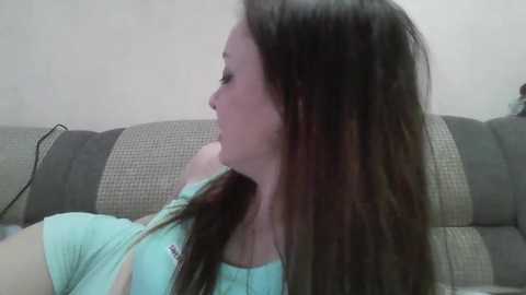 A video of a young woman with long brown hair, wearing a light blue top, lying on a grey and beige woven couch.