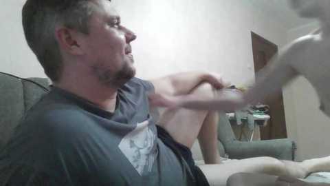 A video of a bearded man in a dark T-shirt, seated in a gray armchair, with a dog playfully licking his face in a cluttered living room.