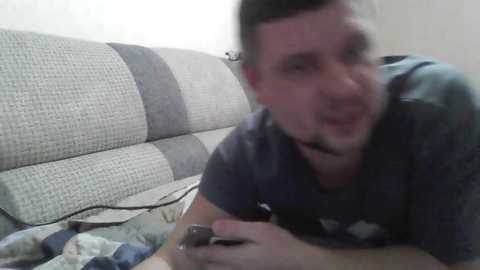 A blurry video of a man with short hair, wearing a headset and a dark T-shirt, playing a video game on a couch with a striped fabric background.