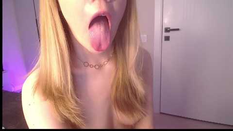 Media: A video of a topless young woman with fair skin and long blonde hair, tongue out, wearing a necklace with \"DOOD\" in a dimly lit room with white walls and a closed door.