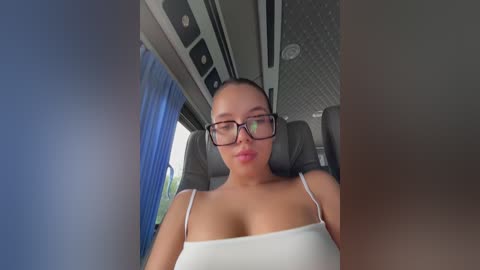Media: Video of a woman with a light skin tone and straight, dark hair in glasses, wearing a white spaghetti-strap top, seated in a modern, well-lit van.