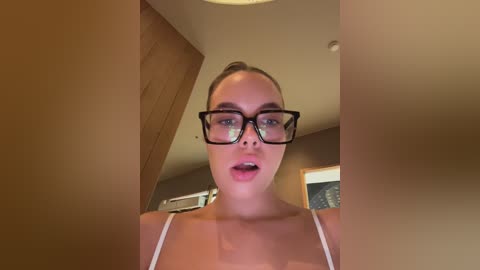 Media: Video of a Caucasian woman with light skin and glasses, wearing a white spaghetti-strap top, standing indoors with wooden beams and a window in the background.