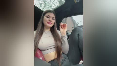 Media: Video of a young Latina woman with long dark hair, wearing a beige crop top and tight black jeans, sitting in a red car.