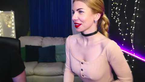 Video of a smiling woman with fair skin and blonde hair in a low bun, wearing a beige cardigan and black choker, standing in a dimly lit living room with blue curtains, fairy lights, and a beige sofa.