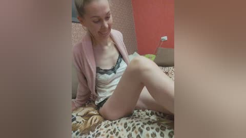 Media: A video of a smiling, fair-skinned woman with blonde hair, wearing a pink cardigan over a black lace bra, sitting on a leopard-print bed, surrounded by a red wall and a beige headboard.