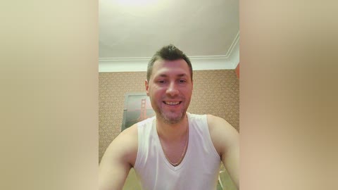 Media: Video of a smiling, muscular, light-skinned man with short dark hair, wearing a white tank top, in a room with beige wallpaper and a framed poster.
