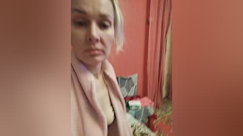 Media: Video of a blonde woman with light skin and a pink robe, standing in a cluttered room with orange walls and a bed covered in clothes.