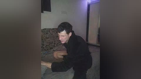 Video of a man with short dark hair, wearing a black coat, kneeling on a gray couch with a patterned cushion, in a dimly lit room with beige walls and a purple light.