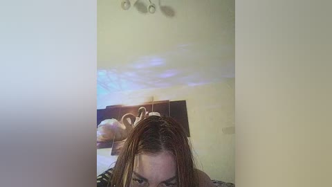 A video shows a young woman with light skin and long, wet brown hair, sitting on a bed with a dark headboard. Her face is partially visible, and she has a playful expression. The room's lighting creates a soft, ambient glow.
