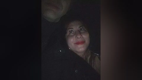 Video of a smiling woman with red lipstick, wearing glasses, and a dark jacket, partially obscured by shadowy figures in the background.