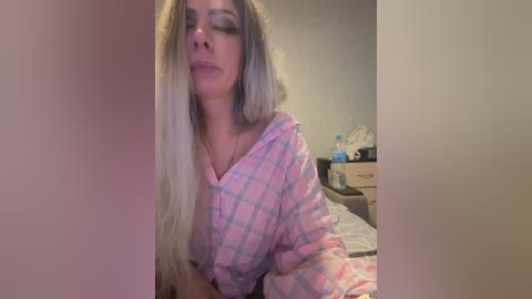 Video of a fair-skinned woman with long, platinum blonde hair wearing a pink and blue plaid pajama shirt. She appears to be in a bedroom with messy surroundings.