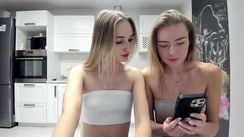 Media: Video of two young blonde women with fair skin in a modern kitchen, wearing strapless white bandeau tops. One woman holds a phone, the other looks at her. White cabinets, black appliances, and graffiti wall art in the background.