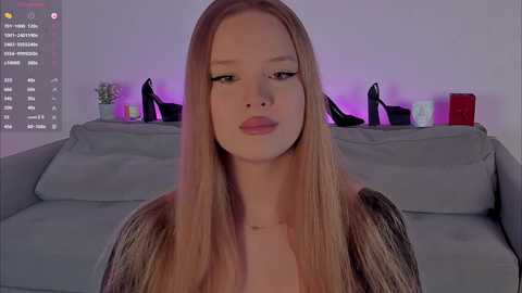 Video of a young woman with long blonde hair, wearing a fur-lined jacket, standing in a modern living room with gray sofa and purple accent lighting.