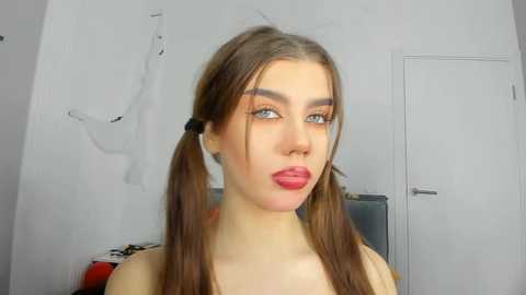 Media: Video of a young, fair-skinned woman with long brown hair in pigtails, wearing red lipstick and a white top. She stands indoors against a plain white wall, with a door and a black and white poster in the background.