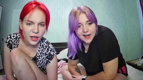 Media: Video of two women with vibrant red and purple hair, smiling, on a bed with leopard-print bedding and a white wall.