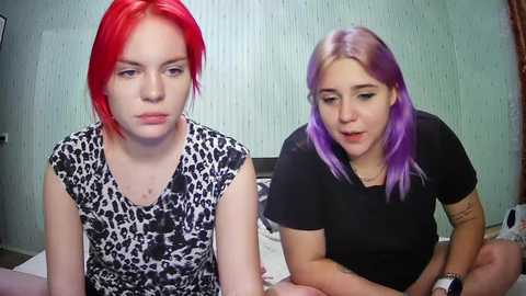 Media: Video of two young women with red and purple hair, seated on a bed. One wears a leopard-print shirt, the other a black T-shirt.