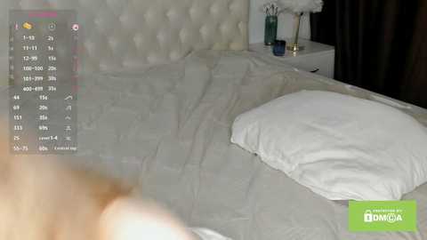 Media: Video of a neatly made bed with white sheets and pillows in a modern bedroom featuring a tufted headboard, a white side table, and a decorative vase.