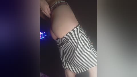 Media: Video of a woman bending over, wearing a black and white striped mini skirt, revealing her bare buttocks. Her skin is light, and she has a bracelet on her wrist. The background is dimly lit, with a blue glow from a TV screen.