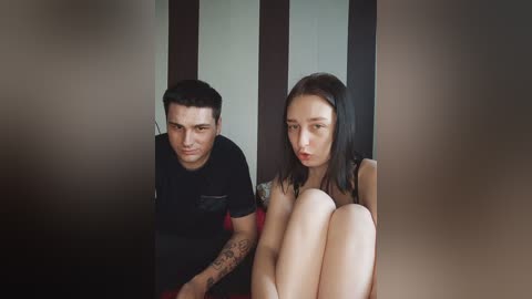 Video of a distressed man with tattoos and a nude woman with fair skin and straight black hair, both sitting on a wooden floor.