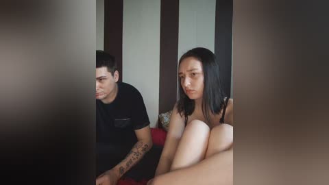Video of a young Asian woman with shoulder-length black hair, sitting nude on a red couch, next to a tattooed man in a black t-shirt.