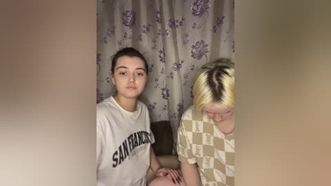 Media: Video of two young women, one with dark hair and a San Francisco State University t-shirt, the other with blonde hair and a checkered shirt, sitting in a dimly lit room with floral-patterned curtains.